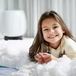 A cheerful young girl happily rests on her bed, with a HomePure air purifier nearby ensuring clean and fresh room air.