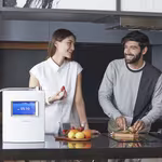 A joyful couple in the kitchen, with a woman ready to enjoy fruit while her husband prepares kiwi with the HomePure Viva Ioniser at the table