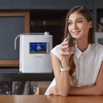 A smiling woman at her kitchen table, holding a glass of HomePure Viva alkaline water, confidently satisfied with her healthy choice.